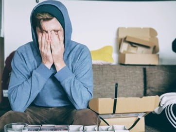Man sitting covering his face in a cluttered room.