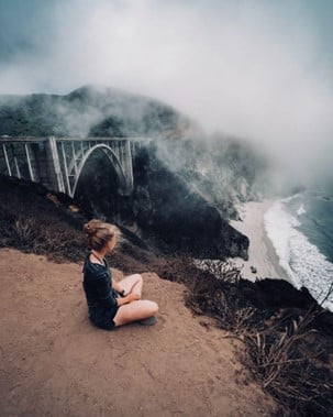 Girl sitting on a sandy hill. Bridge on her left, ocean to her right.