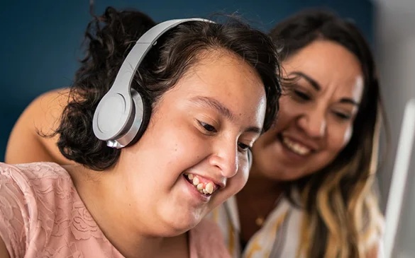 Girl listening to music with headphones while her mom looks on in the background.