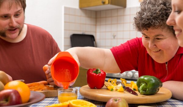 A,Mentally,Disabled,Woman,And,Two,Caretakers,Cooking,Together Group of people sitting around a breakfast table ouring orange juice.